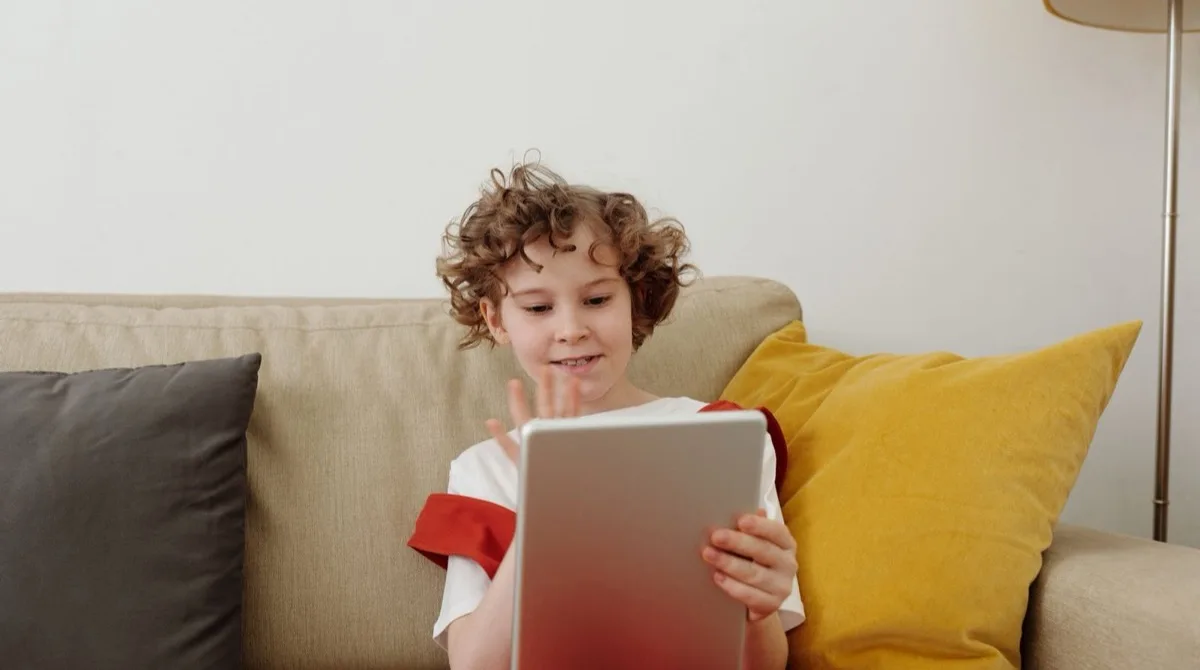 A primary-school-age child sitting on a sofa at home, smiling while practising spelling on a tablet