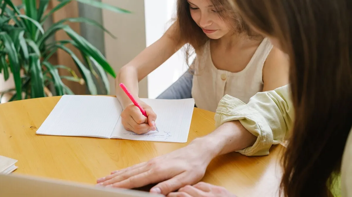 A child writing in a squared exercise book at a wooden table, with a parent sitting beside her